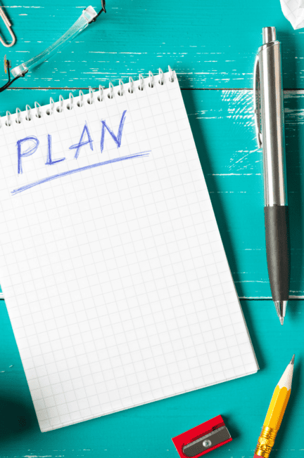 notebook with plan written at the top of a blank page on a desktop with a pen, pencil, and pencil sharpener, and a pair of glasses